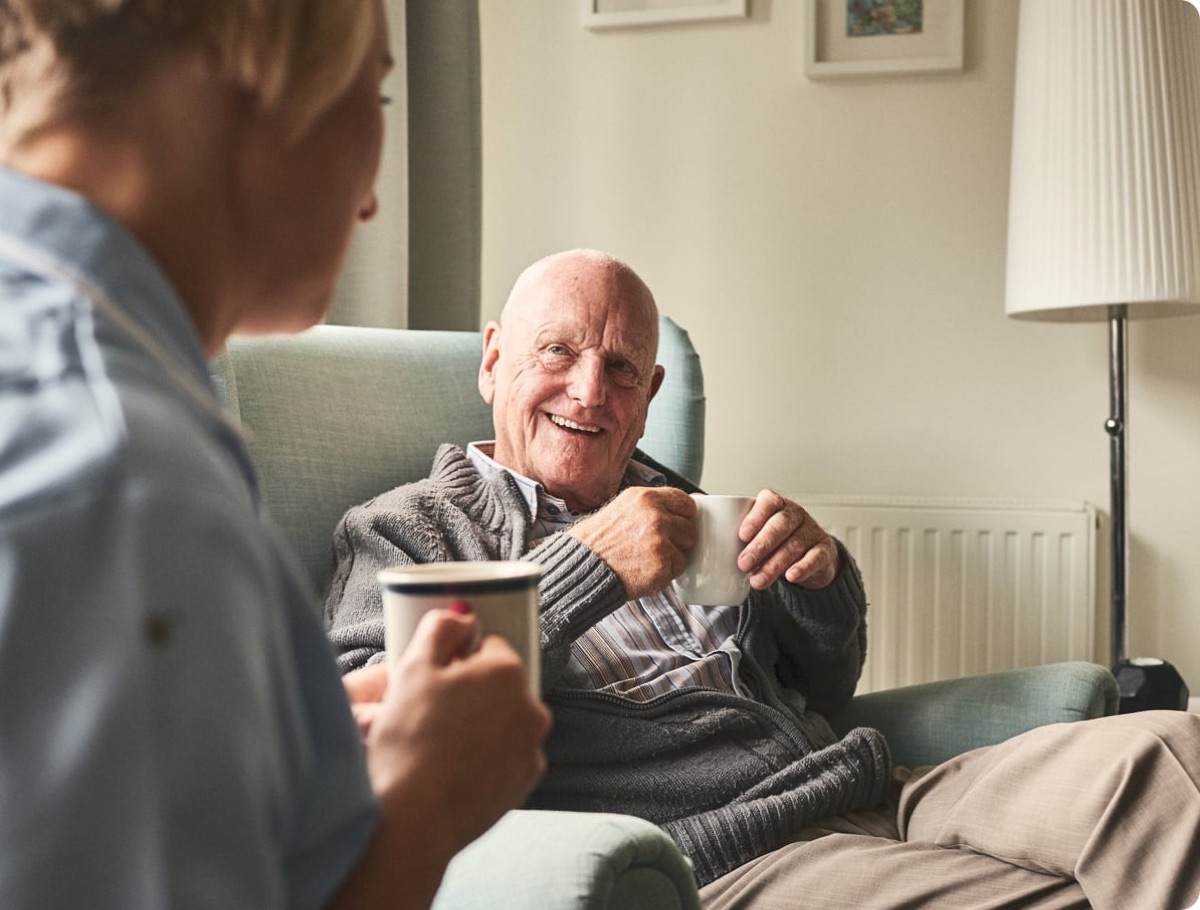 A senior man laughing in his armchair over a mug of coffee with his Caregiver